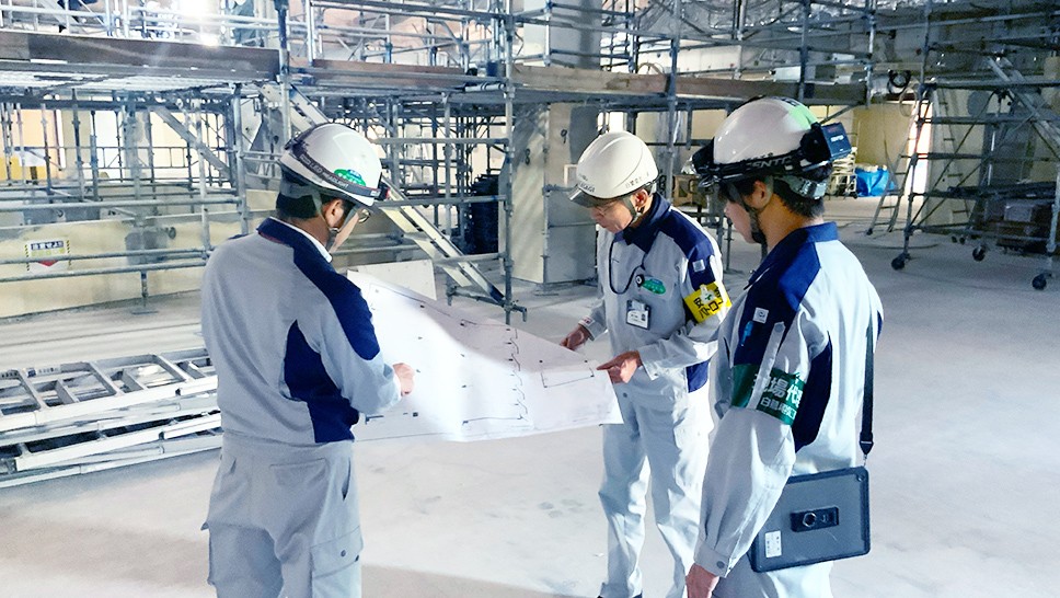 作業服とヘルメットを着用した男性三人が鉄パイプで足場が作られている広い室内で紙を見ている様子の写真。