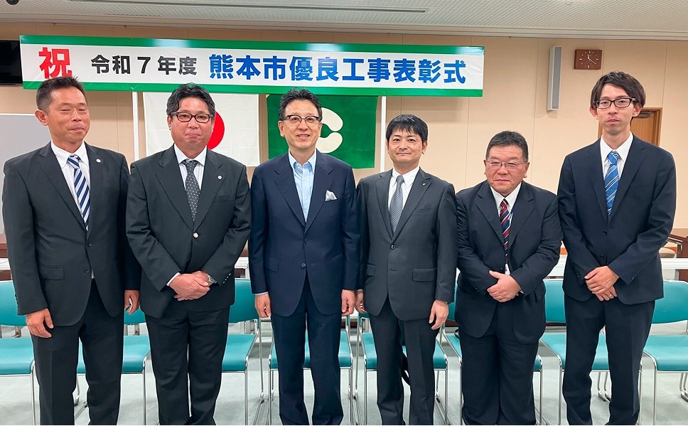 熊本市優良工事表彰式の会場で、スーツ姿の6名の男性が横一列に並んで記念撮影している様子。背後には日本国旗と熊本市の旗、表彰式の横断幕が掲げられている。