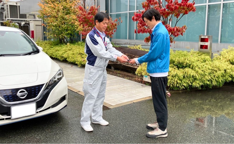 企業の担当者が熊本市へ電気自動車の鍵を渡している様子。左に白い電気自動車、中央に作業服姿の担当者と青い上着の担当者が立っている。