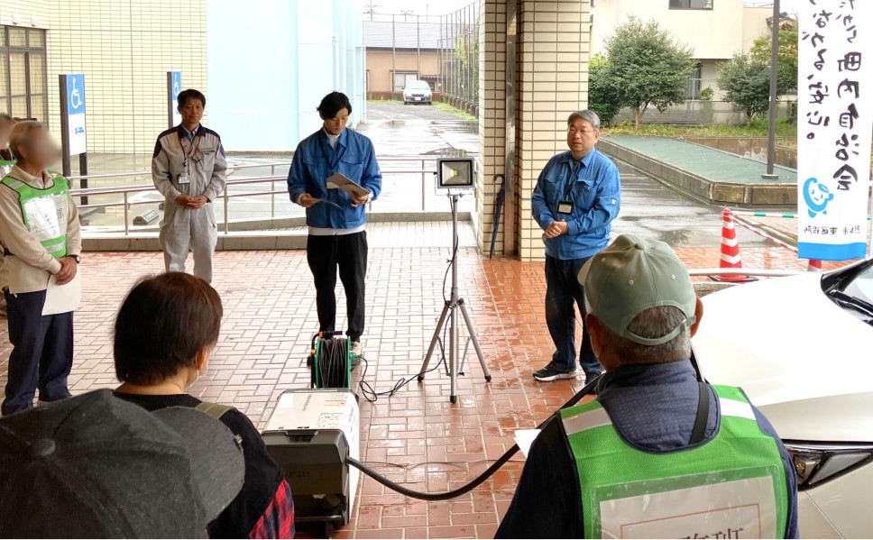 電気自動車からの電力供給訓練の様子。軒下で、参加者が、外部給電器に接続された電気自動車の前で説明を聞いている。