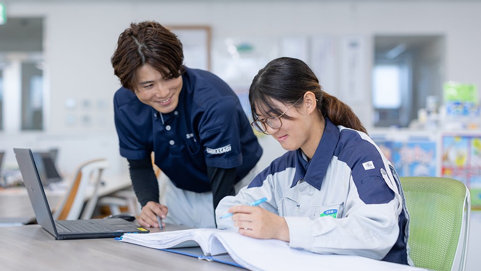 写真：オフィスで女性社員が資料を確認しながら作業し、男性社員が笑顔でサポートしている様子。職場における女性活躍や働きやすい環境づくりを表現した場面。