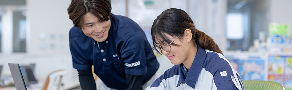 写真：オフィスで女性社員が資料を確認しながら作業し、男性社員が笑顔でサポートしている様子。職場における女性活躍や働きやすい環境づくりを表現した場面。